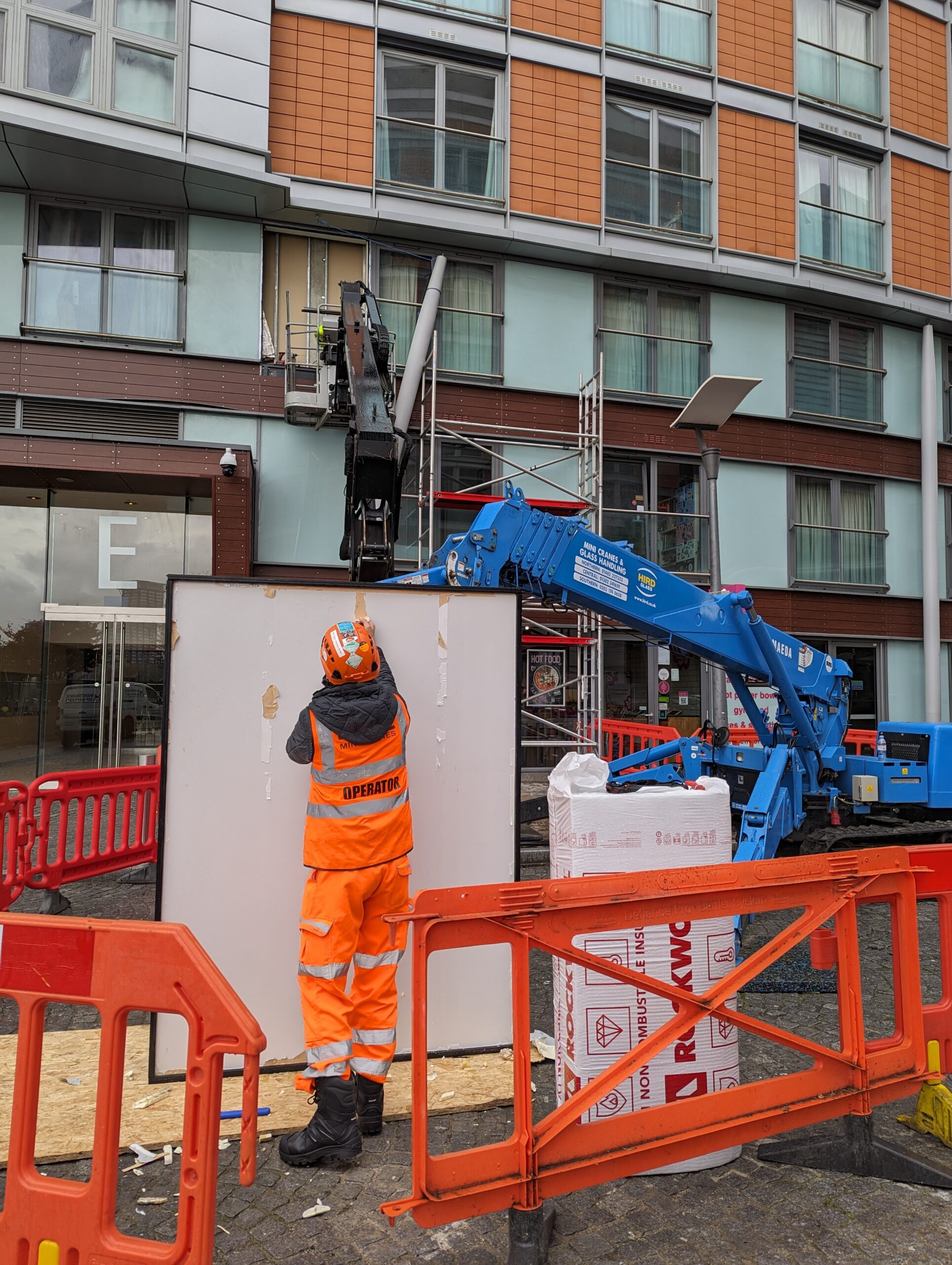A construction worker in an orange uniform stands in front of a building, operating a blue lift. Scaffolding is visible behind them, along with other construction materials. The area is blocked off with red and orange barriers.