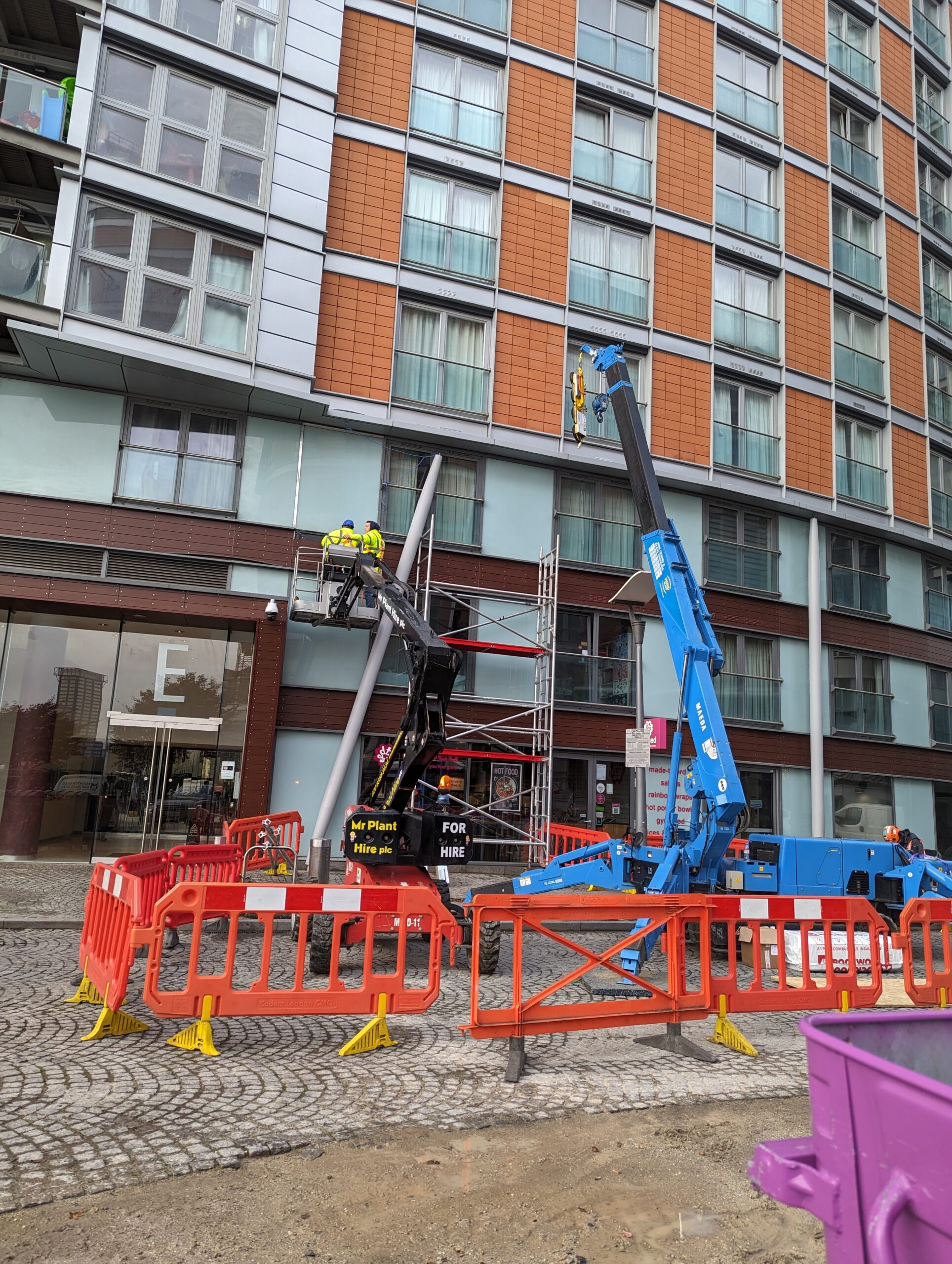 Construction workers in safety gear are elevated on two cherry pickers, one black and one blue, working on the facade of a modern, multi-story building. The area is cordoned off with red and white barriers. Scaffolding is set up between the cherry pickers.