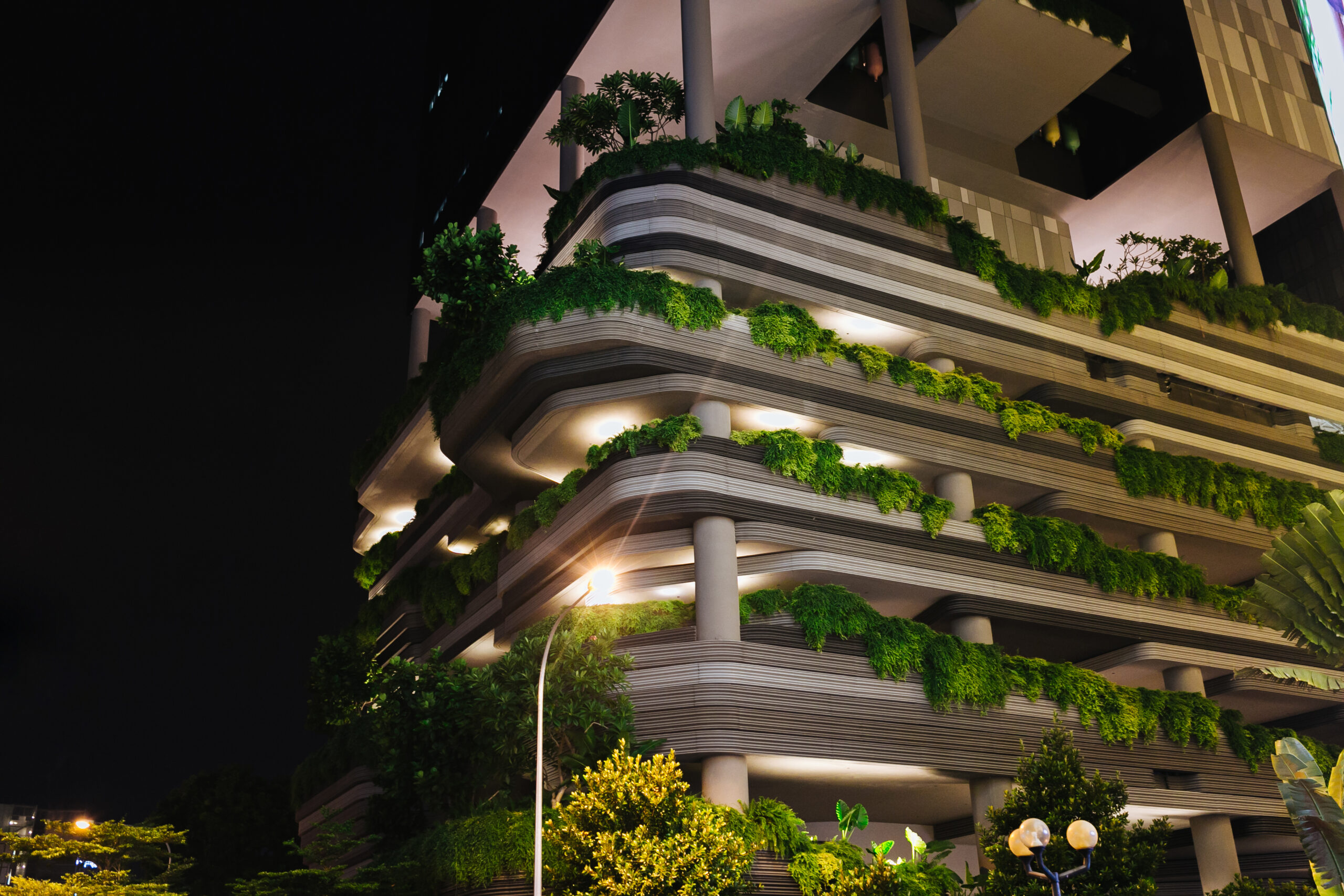 A multi-story building featuring extensive greenery on each level, with lush plants and trees integrated into the green architecture. The structure is illuminated and contrasts with the dark night sky, creating a vibrant urban garden effect.